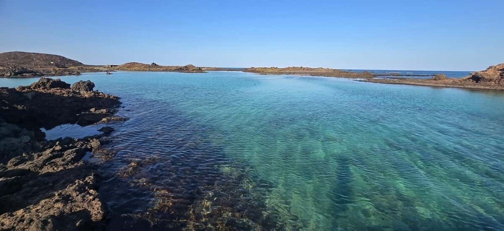 A coastal landscape with clear turquoise waters, rocky shores, and distant hills. 1