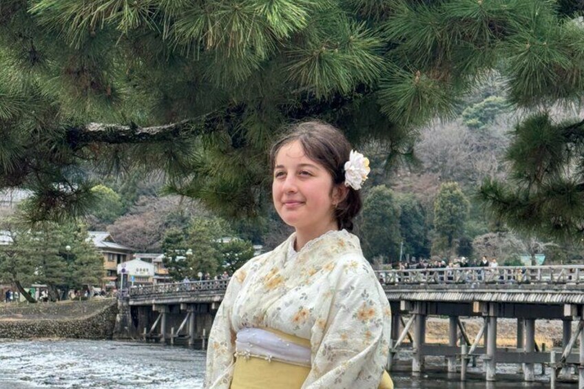 A person in traditional attire standing by a bridge with a backdrop of pine trees and a body of water. 2