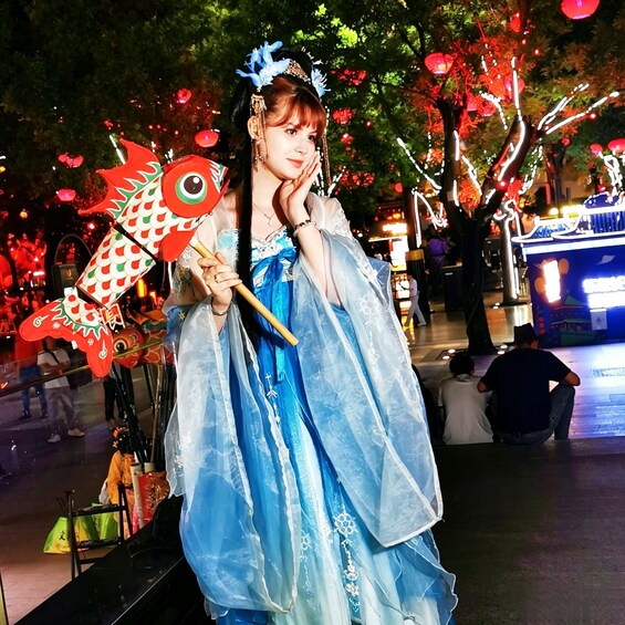 A person in a blue dress holding a red fish-shaped fan, standing in an outdoor area with trees and lights. 5