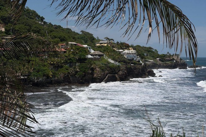 A coastal landscape with waves crashing onto a rocky shore, a lush green hill with buildings, and a clear sky. 2