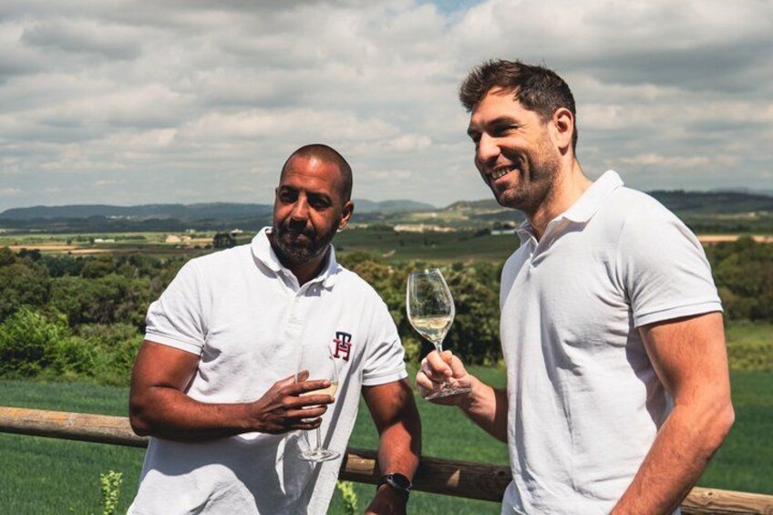 Two men in white polo shirts standing by a wooden fence with a vineyard in the background. 5