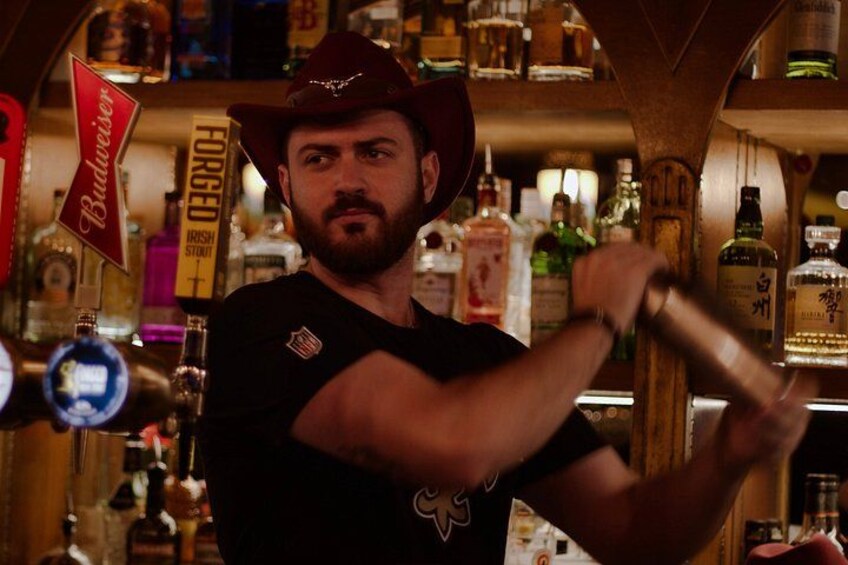 A person wearing a cowboy hat and a black shirt with an American flag emblem, mixing drinks behind a bar with various bottles and a Budweiser sign. 5