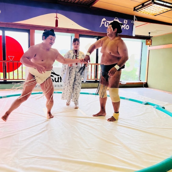 Two sumo wrestlers in a training room with tatami mats, a woman in traditional attire, and a sign with Japanese characters. 4