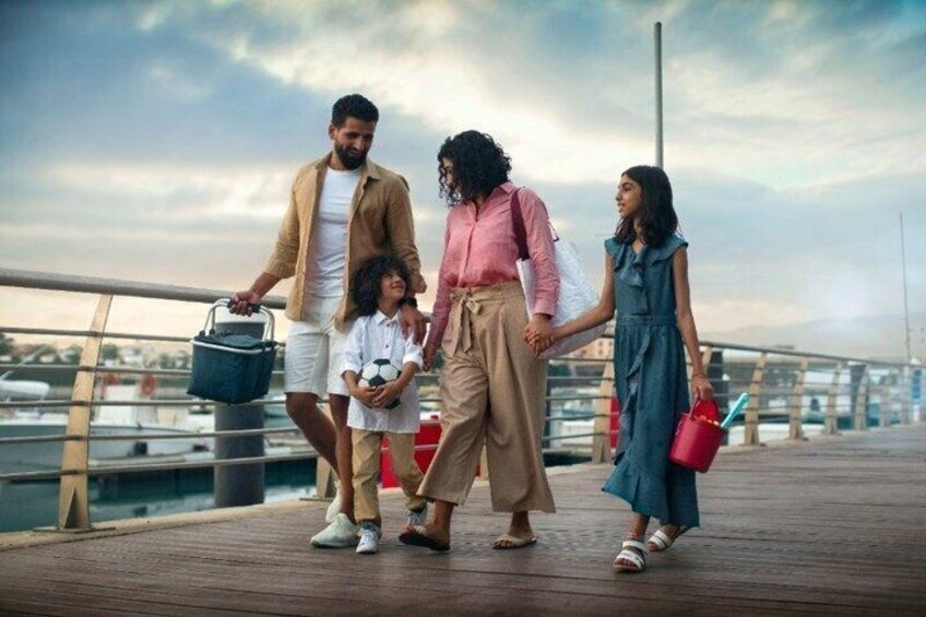 A family walking on a wooden boardwalk by a marina with boats in the background. 1