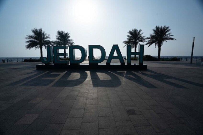 A paved area with large letters spelling 'JEDDAH' and palm trees in the background. 2