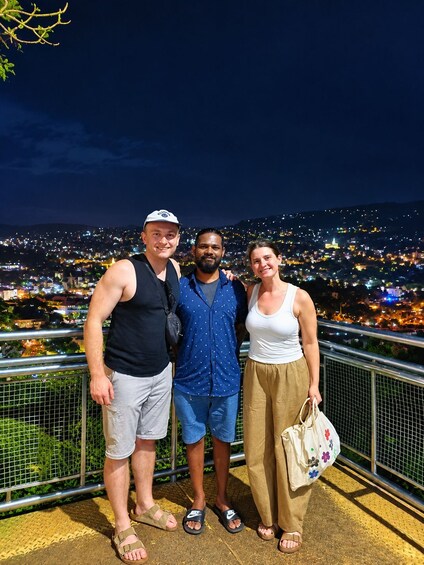 Three people standing on a balcony with a cityscape at night in the background. 4
