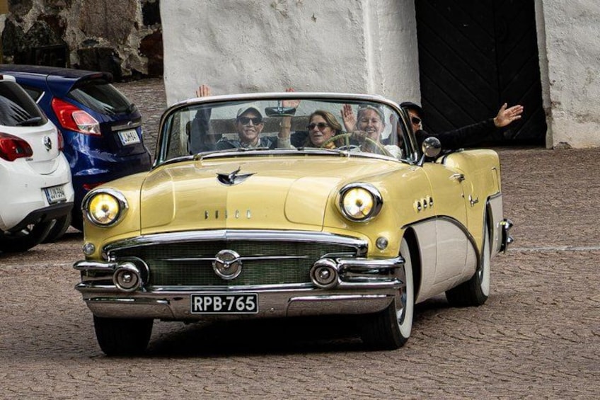 A yellow vintage car with the license plate RPB-765 parked in front of a building with a black door. 1