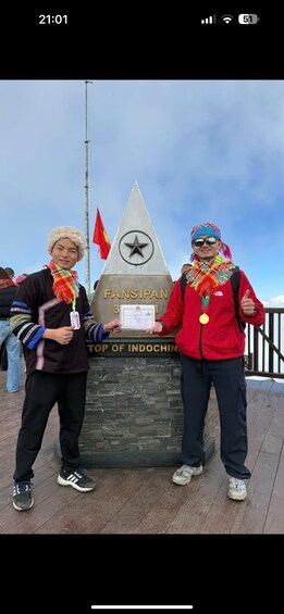 Kaksi ihmistä seisoo monumentin edessä. Monumentissa on teksti: ”Fansipan, Indokiinan kolmas korkein huippu”, ja taustalla näkyy lippu. 4