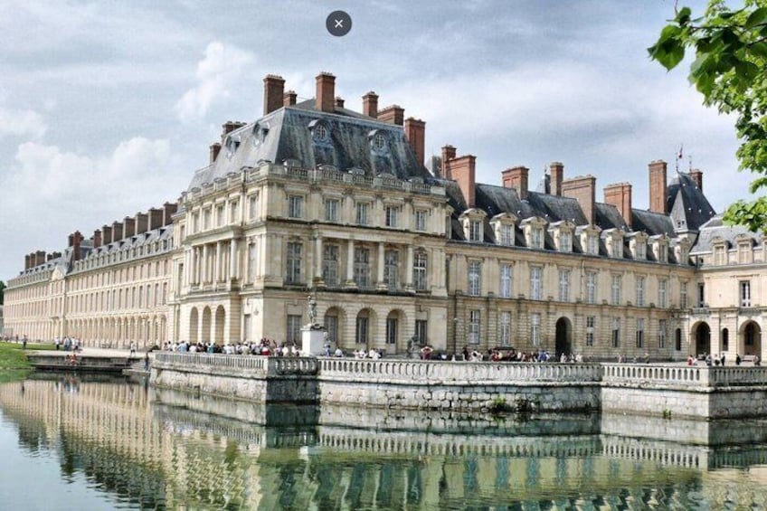 A large historic building with a reflective water body in front, multiple chimneys, and a clear sky. 1