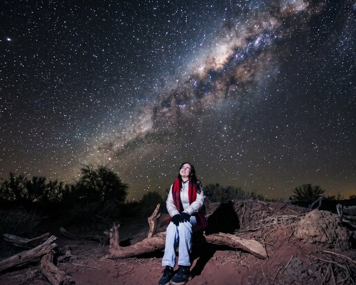 A person sitting on a log under a starry night sky with the Milky Way visible. 1