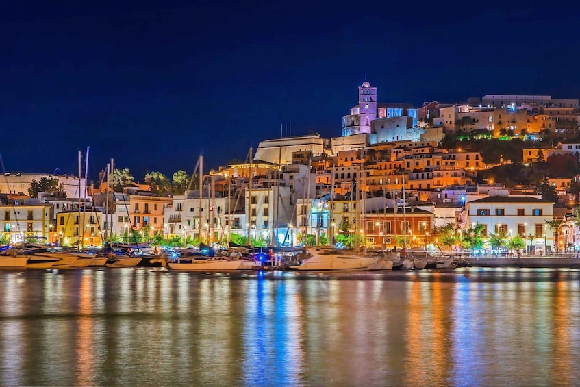 A coastal town at night with illuminated buildings, a prominent tower, and boats docked along the waterfront. 4