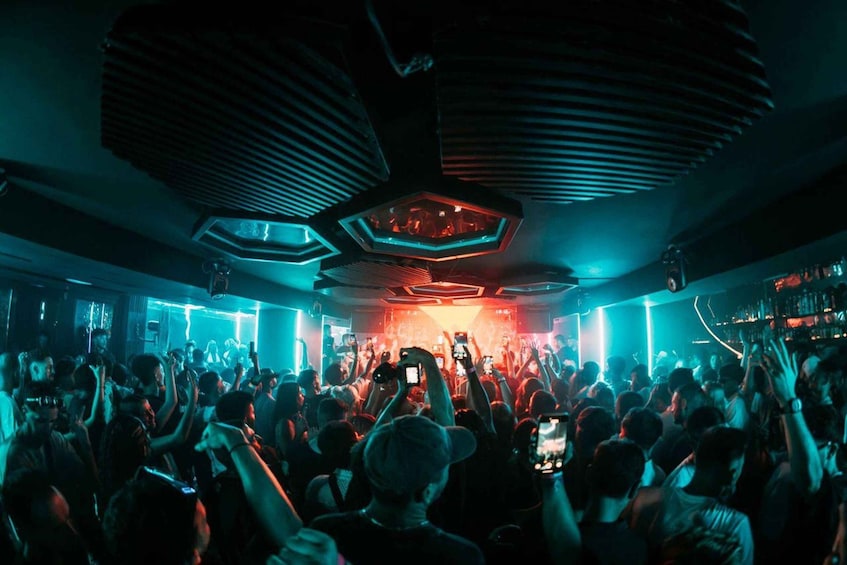 A crowded indoor venue with a stage, geometric ceiling design, and ambient lighting. 1