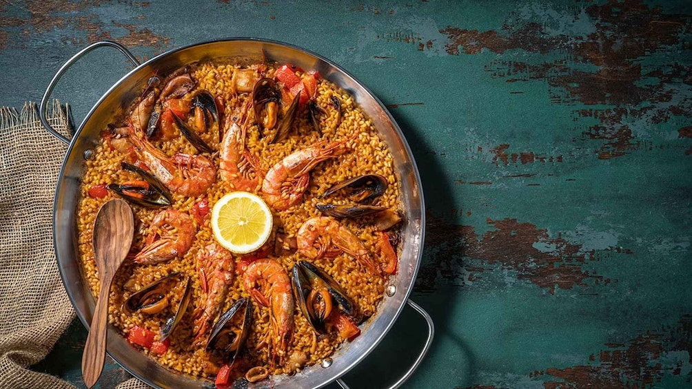 A large pan of seafood paella with mussels and shrimp, a lemon slice, and a wooden spoon on a rustic table. 5
