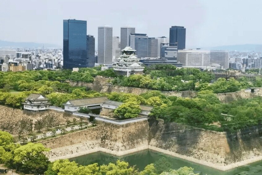Perinteinen japanilainen linna, jonka ympärillä on vehreää kasvillisuutta, ja taustalla näkyy moderneja pilvenpiirtäjiä. 1