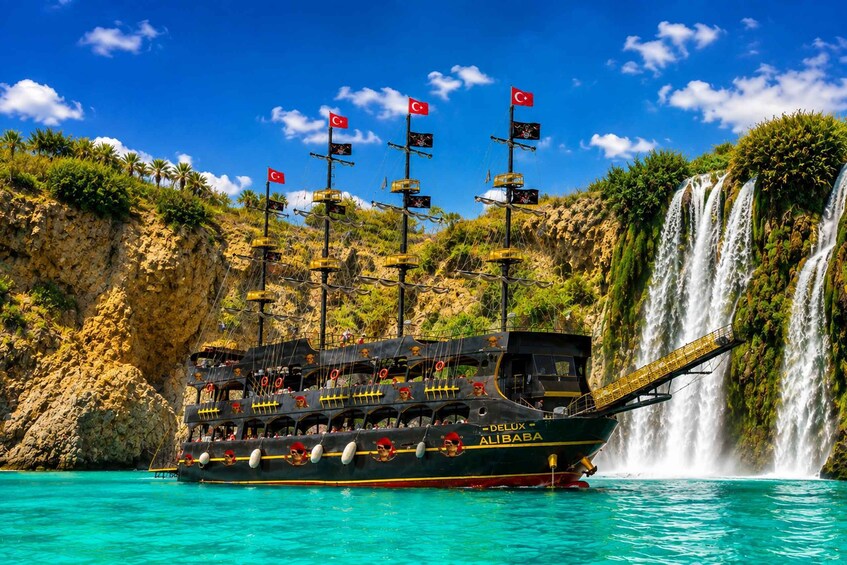 A pirate-themed boat named DELUX ALIBABA on turquoise waters with a waterfall in the background. 1