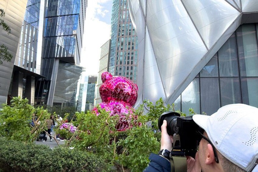 A person taking a photo of a pink bear sculpture in an urban setting with modern buildings in the background. 3