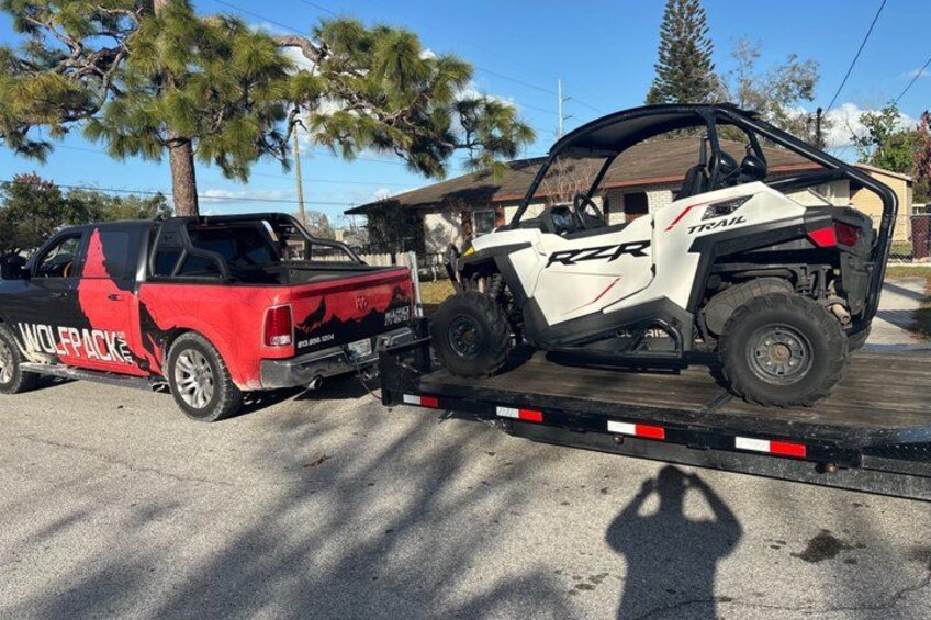 A white RZR on a flatbed trailer, a red truck with 'WOLF PACK' written on it, and a residential area with trees and houses in the background. 3