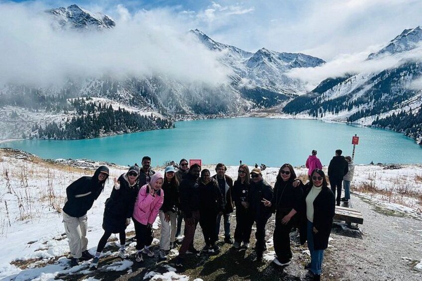 A group of people standing on a snowy mountain overlooking a turquoise lake with snow-capped mountains in the background. 1