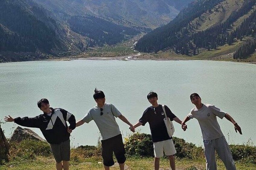 Four people standing on a grassy area overlooking a lake with mountains in the background. 2