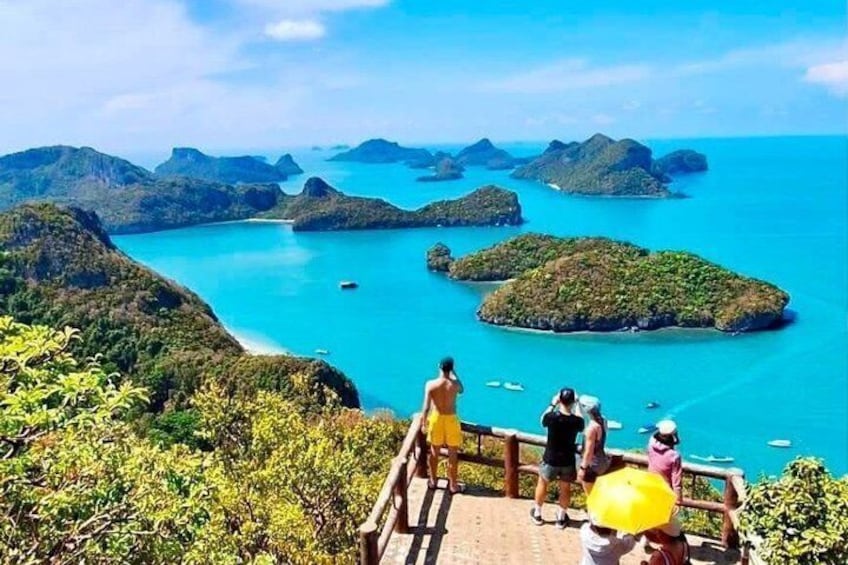 A coastal landscape with a wooden viewing platform, lush greenery, and turquoise waters with boats. 3
