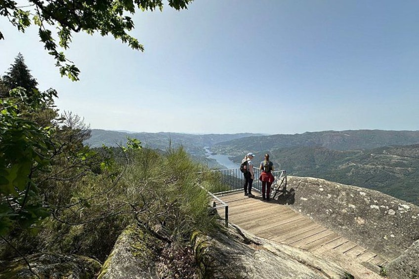 A wooden observation deck with two people standing on it, overlooking a valley with a river and distant mountains. 1