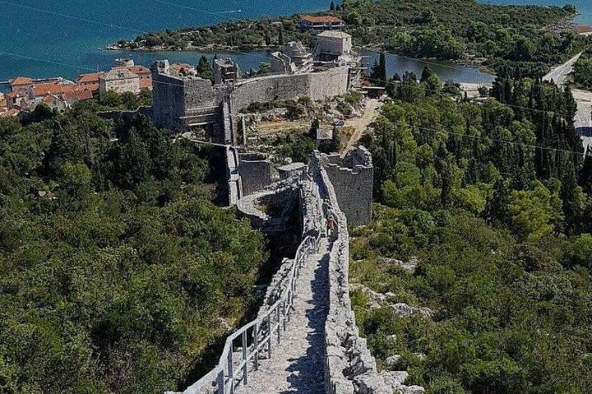 A historical stone wall leading to a fortified structure with a view of a body of water and surrounding greenery. 5