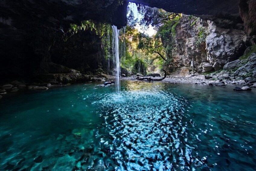 A clear blue pool with a waterfall, surrounded by rocky cliffs and greenery. 1