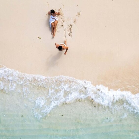A sandy beach with two people sunbathing and a gentle wave approaching. 1