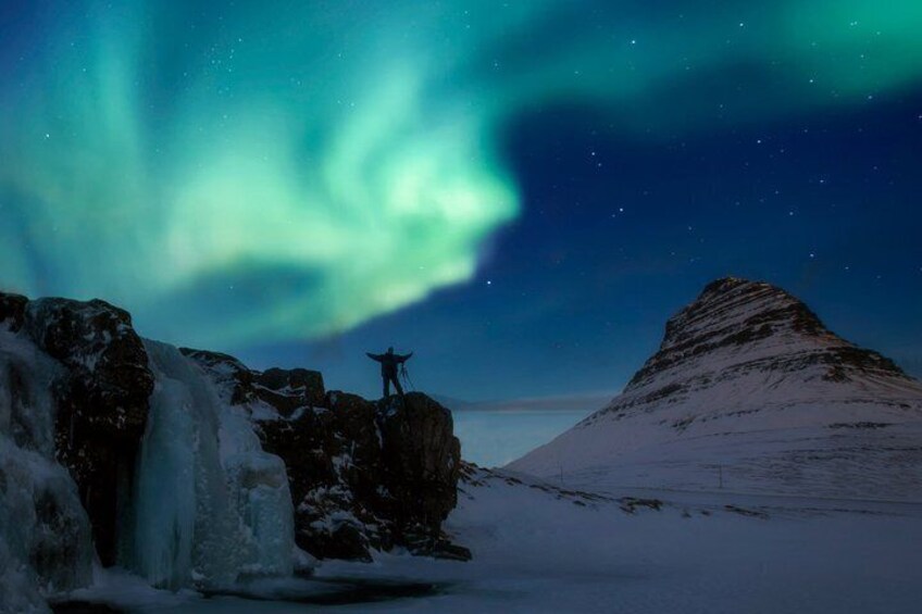 A person standing on a snowy hill with a waterfall in the foreground and the aurora borealis in the background. 1