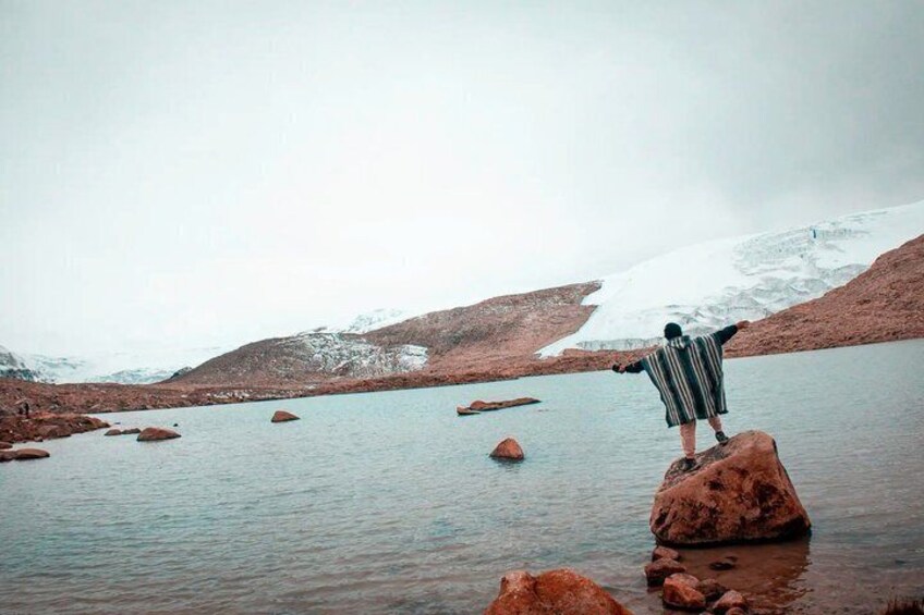 A person standing on a rock in a body of water with snow-capped mountains in the background. 1
