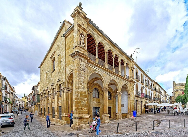 A historic stone building with arches and a balcony, surrounded by cobblestone streets and pedestrians. 5
