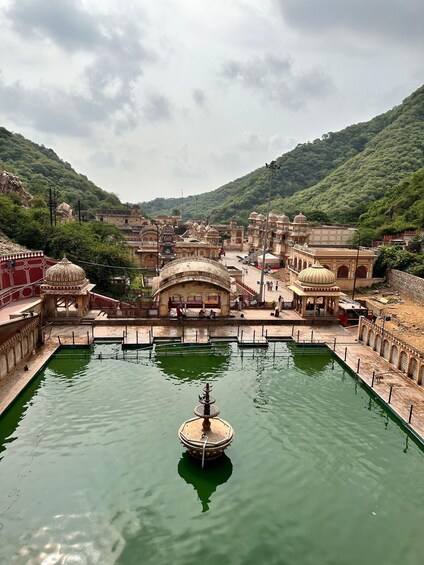 A historic square with a central water fountain, surrounded by traditional architecture and lush green hills in the background. 2