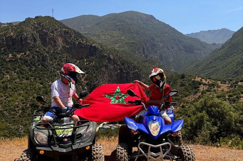 Two people on ATVs with a flag between them, surrounded by mountainous terrain. 5