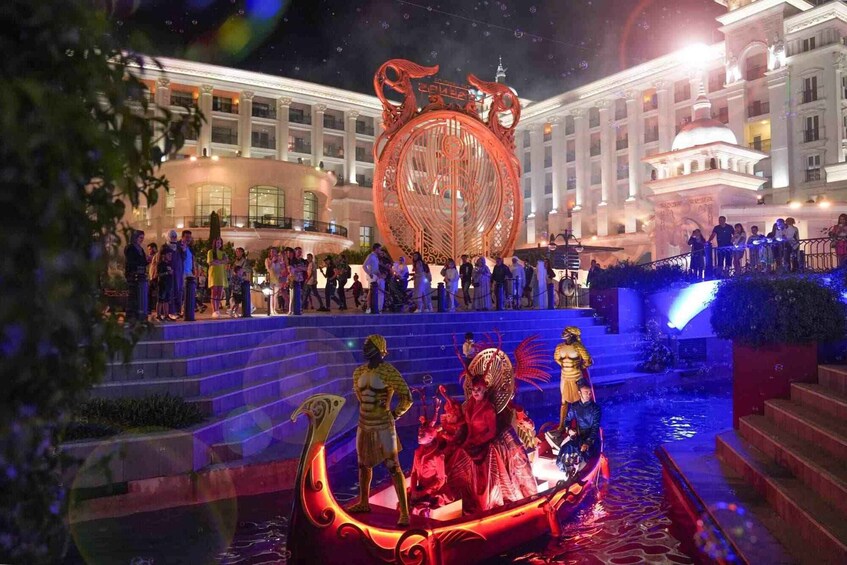 A nighttime outdoor event with a large illuminated circular structure, a water feature with figures, and a building in the background. 5