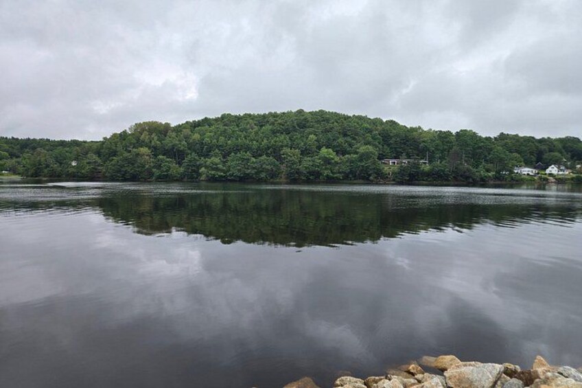 A calm lake with a forested hill in the background and a rocky shoreline in the foreground. 4
