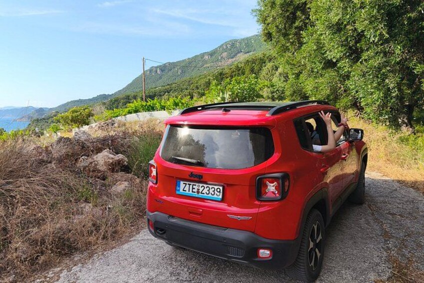 A red SUV parked on a gravel path with a scenic view of a coastal landscape and greenery in the background. 3