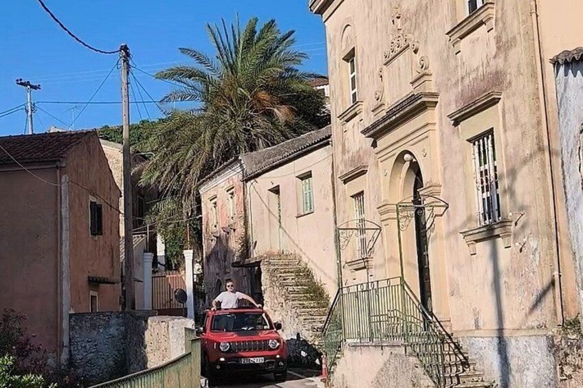 A narrow street with a red car parked on the side, a staircase leading to a building, and palm trees in the background. 2