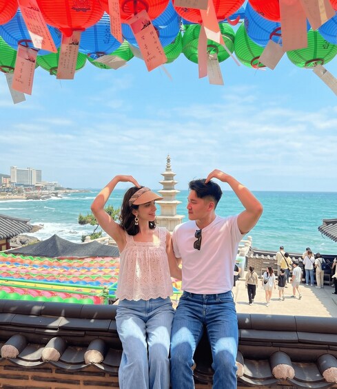 A couple making a heart shape with their hands, overlooking a coastal area with a pagoda and colorful umbrellas. 3