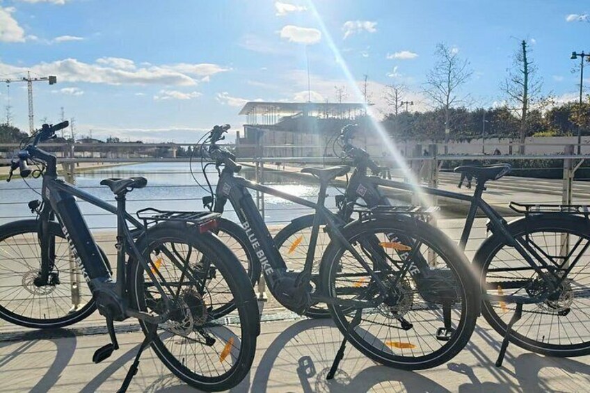 Three bicycles parked near a waterfront with a bridge and buildings in the background. 1