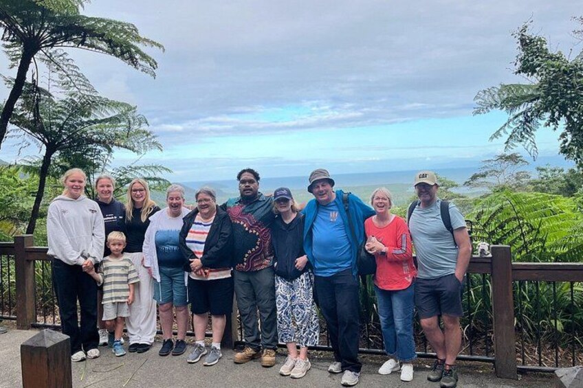 A group of people standing on a wooden platform with a scenic view of a forested landscape and a body of water in the distance. 1