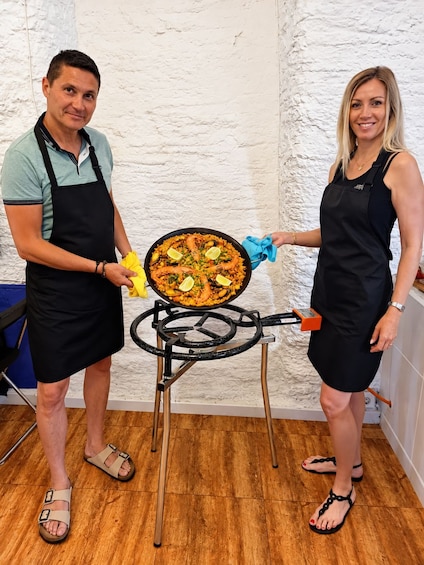 Two people holding a large pan of food, standing in a kitchen with a white textured wall and wooden floor. 3