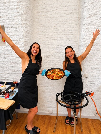 Two people in black aprons holding a pan of food, with a grill in the background. 1