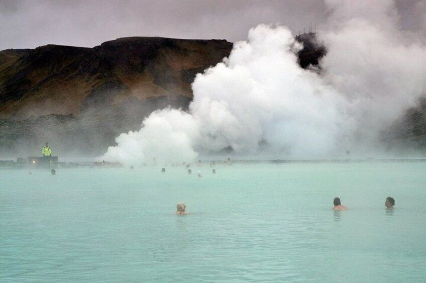 A geothermal spa with people swimming in clear water, steam rising from the ground, and a person in a high-visibility jacket overseeing the area. 4