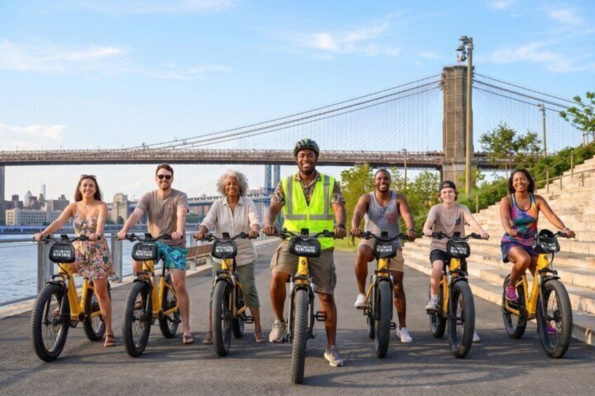 A group of people on bicycles by a waterfront with a bridge in the background. 2