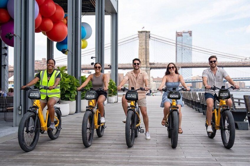 Four people riding electric bikes on a wooden boardwalk with a bridge in the background. 4