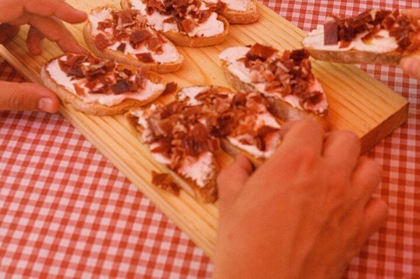 A wooden board with several pieces of toast topped with a white spread and brown toppings, placed on a red and white checkered tablecloth. 1