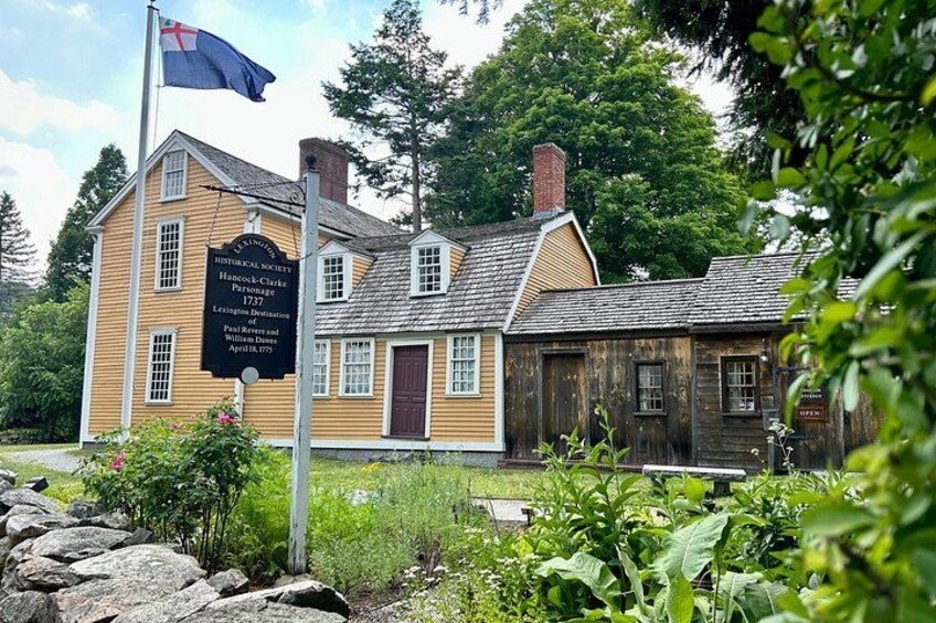 A historical house with a signboard, surrounded by greenery and a stone wall. 2