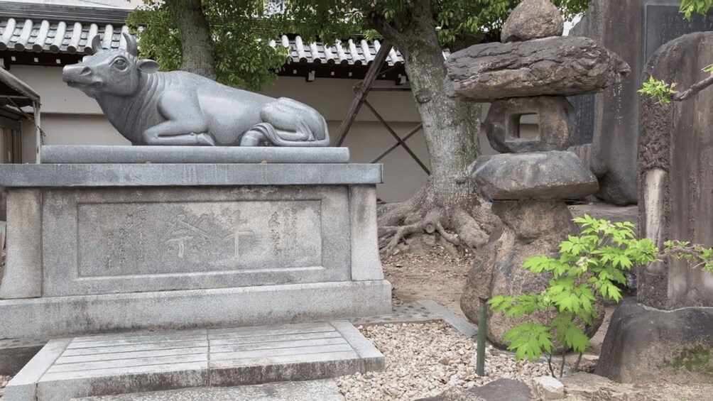 A stone sculpture of a cow on a pedestal, a tree, and a rock formation with inscriptions. 2