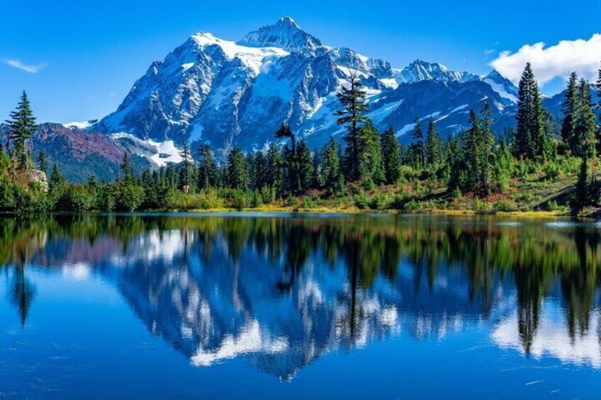 A mountain range with snow-capped peaks, a calm lake reflecting the scenery, and dense forested area. 1