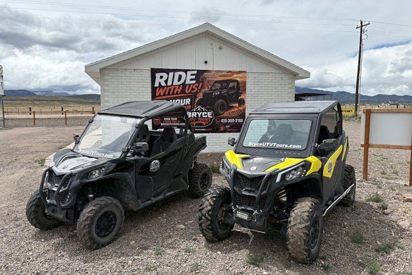 Two off-road vehicles parked in front of a building with a sign that reads 'RIDE WITH US'. 1